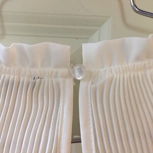 May include: White pleated fabric with a white button closure. The fabric is gathered at the top and bottom.