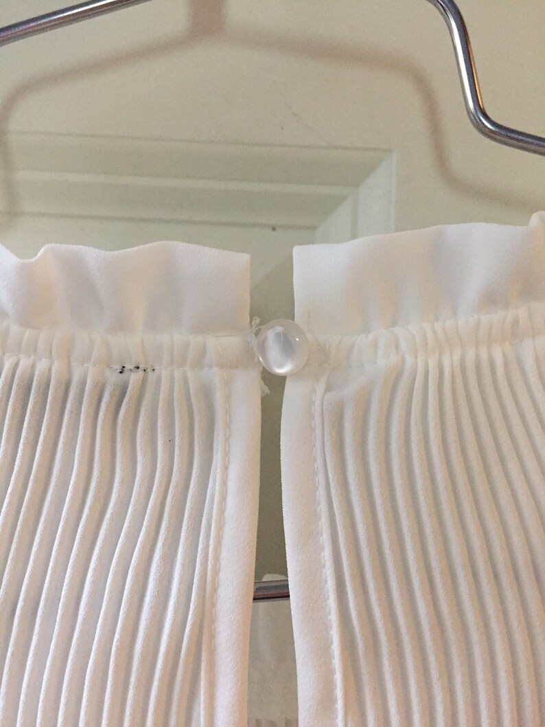 May include: White pleated fabric with a white button closure. The fabric is gathered at the top and bottom.