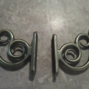 May include: Two matching metal wall brackets with a brushed antique brass finish. Each bracket features a curved, scroll-like design with multiple circular loops and a flat, vertical mounting plate with pre-drilled holes for installation.