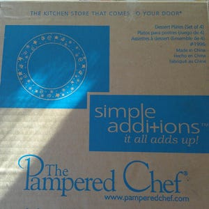 May include: A cardboard box featuring a blue square with a circular design of stars and dots. The box also has the text "simple additions it all adds up!" and "The Pampered Chef" logo. The box contains dessert plates.
