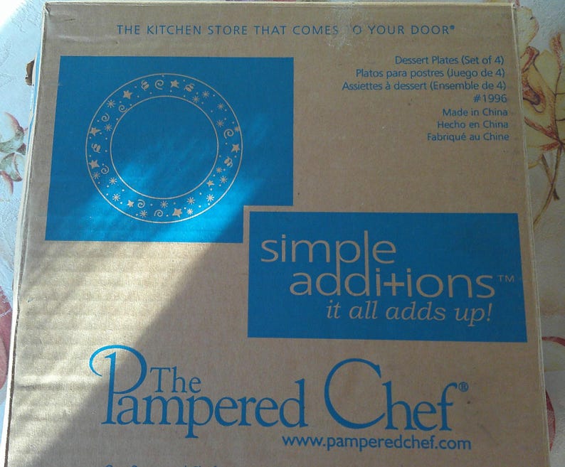 May include: A cardboard box featuring a blue square with a circular design of stars and dots. The box also has the text "simple additions it all adds up!" and "The Pampered Chef" logo. The box contains dessert plates.