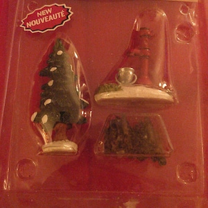 May include: A set of three miniature Christmas village decorations: a water pump, a tree, and a pile of firewood. The set is packaged in a clear plastic clamshell with a red background. The packaging includes the text "WATER PUMP, TREE & FIREWOOD, Set of 3" and "POMPE À EAU, ARBRE ET BOIS DE CHAUFFAGE, Jeu de 3".