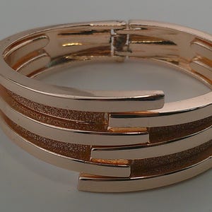 May include: A rose gold bracelet with a hinged clasp. The bracelet features a layered design with three textured bands and two smooth bands. The jewelry is a stylish accessory.