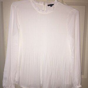 May include: White long-sleeved blouse with a ruffled collar and pleated detailing.