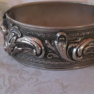 May include: Silver-toned hinged bangle bracelet with ornate floral and leaf detailing. The bracelet features raised relief designs of stylized leaves and flowers, with a textured finish. A classic piece of jewelry.