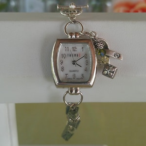 May include: Silver-toned quartz watch with a rectangular face. The watch has a white dial with black numerals and hands. Decorative charms dangle from the watch. The watch band is a green, textured material.