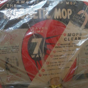 May include: A packaged vintage "The American Magnetic Mop" with red and black accents. The packaging includes text about the mop's features, such as saving time and cleaning floors. The package is sealed in clear plastic.