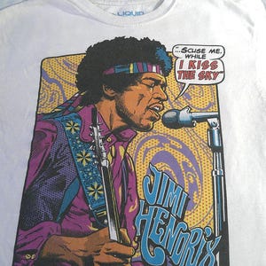 May include: White t-shirt featuring a graphic of Jimi Hendrix playing guitar and singing into a microphone. The image includes the text "...Scuse me while I kiss the sky" in a speech bubble, and "Jimi Hendrix" in blue letters.