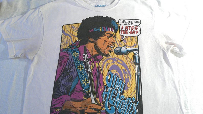 May include: White t-shirt featuring a graphic of Jimi Hendrix playing guitar and singing into a microphone. The image includes the text "...Scuse me while I kiss the sky" in a speech bubble, and "Jimi Hendrix" in blue letters.