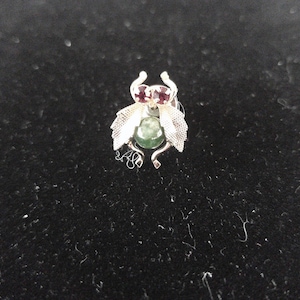 May include: A silver brooch with a green stone and two red stones. The brooch is shaped like a stylized insect with wings.
