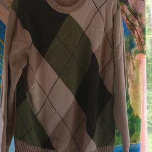 May include: A beige argyle sweater with a crew neck and long sleeves. The argyle pattern features diamonds in shades of green, black, and beige. The sweater is hanging on a black hanger.