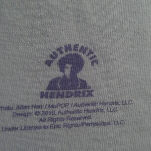 May include: Close-up of a light blue fabric with a purple graphic. The graphic features the word "AUTHENTIC" above a silhouette of a person and the word "HENDRIX" below. Text at the bottom includes copyright information.