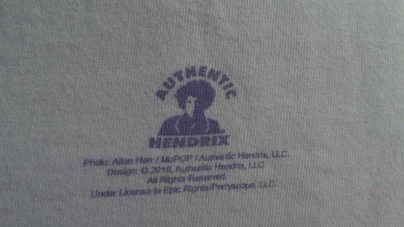 May include: Close-up of a light blue fabric with a purple graphic. The graphic features the word "AUTHENTIC" above a silhouette of a person and the word "HENDRIX" below. Text at the bottom includes copyright information.