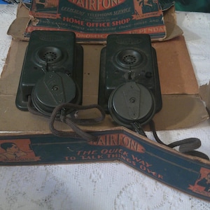 May include: Two vintage Fairfone telephones with rotary dials and dark green housings. The phones are displayed with their original packaging, which includes printed text and illustrations. The packaging also includes the text "The Quick Way to Talk Things Over."