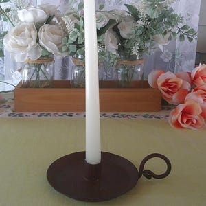 May include: A tall, slender white taper candle stands upright in a dark brown, circular metal candle holder. The holder features a small, curved handle on one side, providing a simple and functional design for holding the candle securely in place.