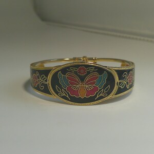 May include: Gold-toned hinged bracelet with a black enamel band. The bracelet features a central oval design with a butterfly motif in red, blue, and yellow, surrounded by floral accents. The bracelet has a secure clasp.