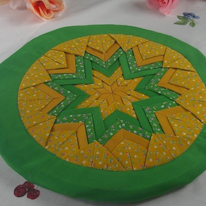 May include: A circular fabric mat featuring a folded star quilt pattern in shades of yellow, green, and white. The center displays a multi-pointed star design created with layered fabric triangles, surrounded by a solid green fabric border.