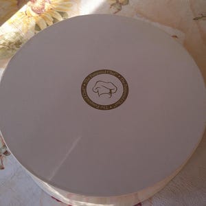 May include: A round, off-white cardboard box with a lid. The center of the lid features a circular logo with the text "The Pampered Chef" surrounding a chef's hat icon. The box is likely for food storage or transport.
