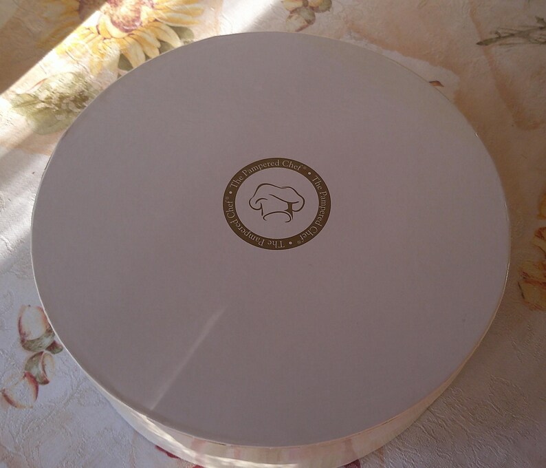 May include: A round, off-white cardboard box with a lid. The center of the lid features a circular logo with the text "The Pampered Chef" surrounding a chef's hat icon. The box is likely for food storage or transport.