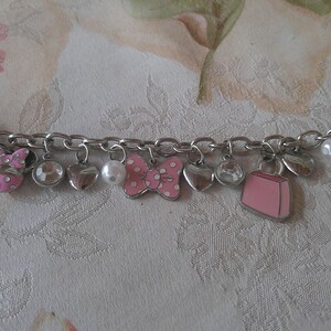 Puede incluir: Pulsera de abalorios plateada con varios dijes, incluyendo corazones, lazos y un bolso rosa. Los dijes est&aacute;n acentuados con esmalte rosa y pedrer&iacute;a transparente. Tambi&eacute;n se incluye una perla blanca. La pulsera est&aacute; sobre un fondo floral.