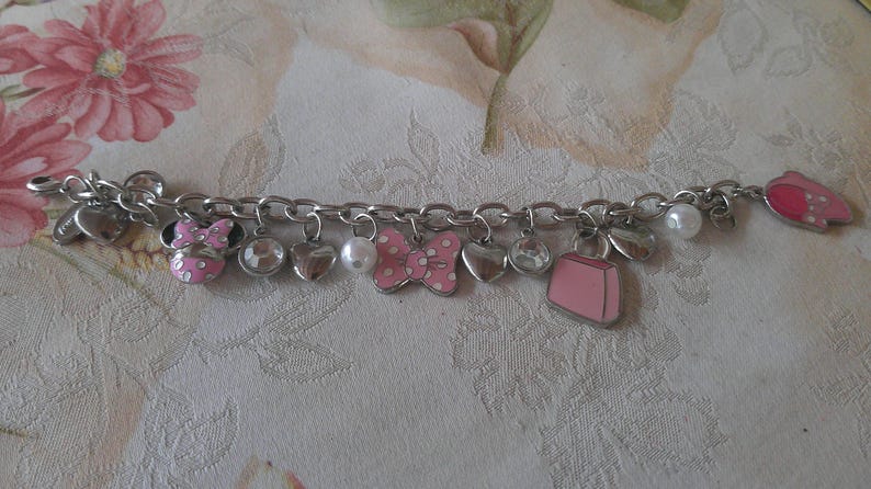 Puede incluir: Pulsera de abalorios plateada con varios dijes, incluyendo corazones, lazos y un bolso rosa. Los dijes est&aacute;n acentuados con esmalte rosa y pedrer&iacute;a transparente. Tambi&eacute;n se incluye una perla blanca. La pulsera est&aacute; sobre un fondo floral.