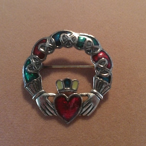 May include: A silver-toned Claddagh brooch with a red heart, a crown, and hands. The circular design features a colorful enamel band in red, green, and blue. The brooch is a traditional Irish symbol of love, loyalty, and friendship.