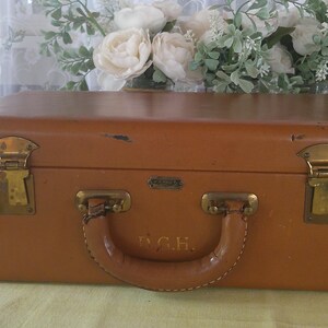 May include: A rectangular tan hard-shell suitcase featuring brass-toned metal latches, a central handle with the embossed initials D.G.H., and a small rectangular metal plate centered above the handle. The exterior shows minor surface wear.