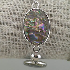 May include: Silver-toned bracelet featuring an oval abalone shell pendant. The iridescent shell displays a spectrum of colors, including blues, greens, and purples. The bracelet has a toggle clasp and a heart-shaped charm.