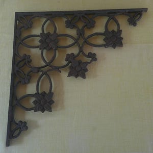 May include: A dark brown, L-shaped metal shelf bracket featuring an intricate, openwork design of interconnected loops and stylized floral motifs. The bracket has a flat top and side edge, with a repeating pattern of four-petaled flower shapes.