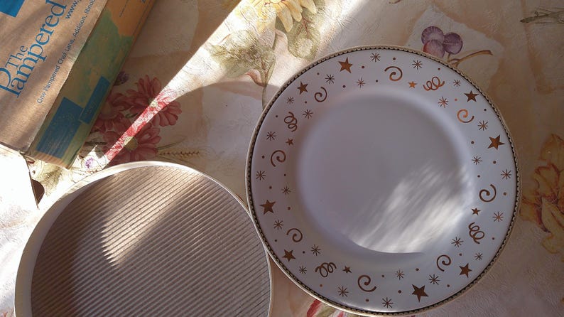 May include: White ceramic plate with a gold rim and festive star and swirl design. A second, light-colored, ridged plate is also visible. The items are on a floral patterned surface, with a box in the background.