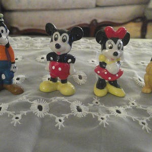 May include: A set of five ceramic Disney character figurines. The figurines are Mickey Mouse, Minnie Mouse, Goofy, Pluto, and Donald Duck. The figurines are all in a standing position and are painted in their signature colors.