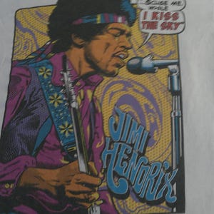 May include: White t-shirt featuring a colorful graphic of Jimi Hendrix playing a guitar and singing into a microphone. The design includes the text "I Kiss The Sky" and "Jimi Hendrix".