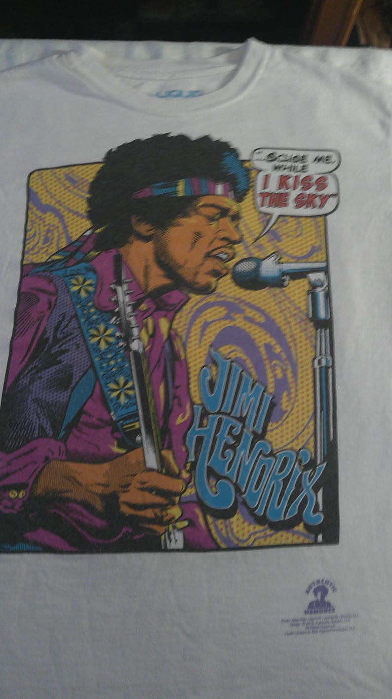 May include: White t-shirt featuring a colorful graphic of Jimi Hendrix playing a guitar and singing into a microphone. The design includes the text "I Kiss The Sky" and "Jimi Hendrix".