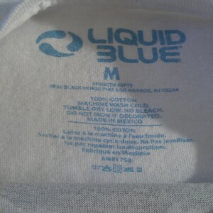 May include: Close-up of a white t-shirt tag with blue text. The tag features the "Liquid Blue" logo, size "M", and washing instructions. The tag also indicates the shirt is 100% cotton and made in Mexico.