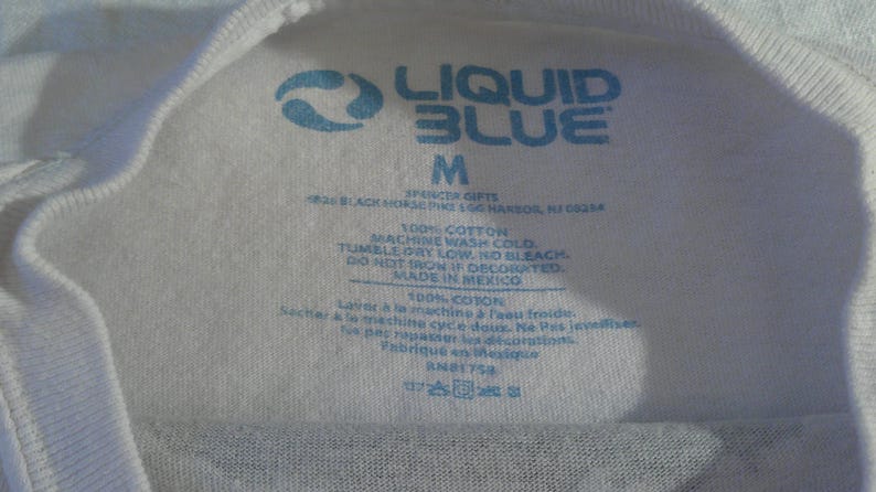 May include: Close-up of a white t-shirt tag with blue text. The tag features the "Liquid Blue" logo, size "M", and washing instructions. The tag also indicates the shirt is 100% cotton and made in Mexico.