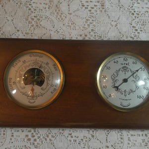 May include: A rectangular wooden wall plaque featuring two circular brass-rimmed gauges. The left gauge is a barometer labeled Stormy, Rain, Change, Fair, and Dry. The right gauge is a hygrometer labeled % rel. with a scale from 0 to 100.