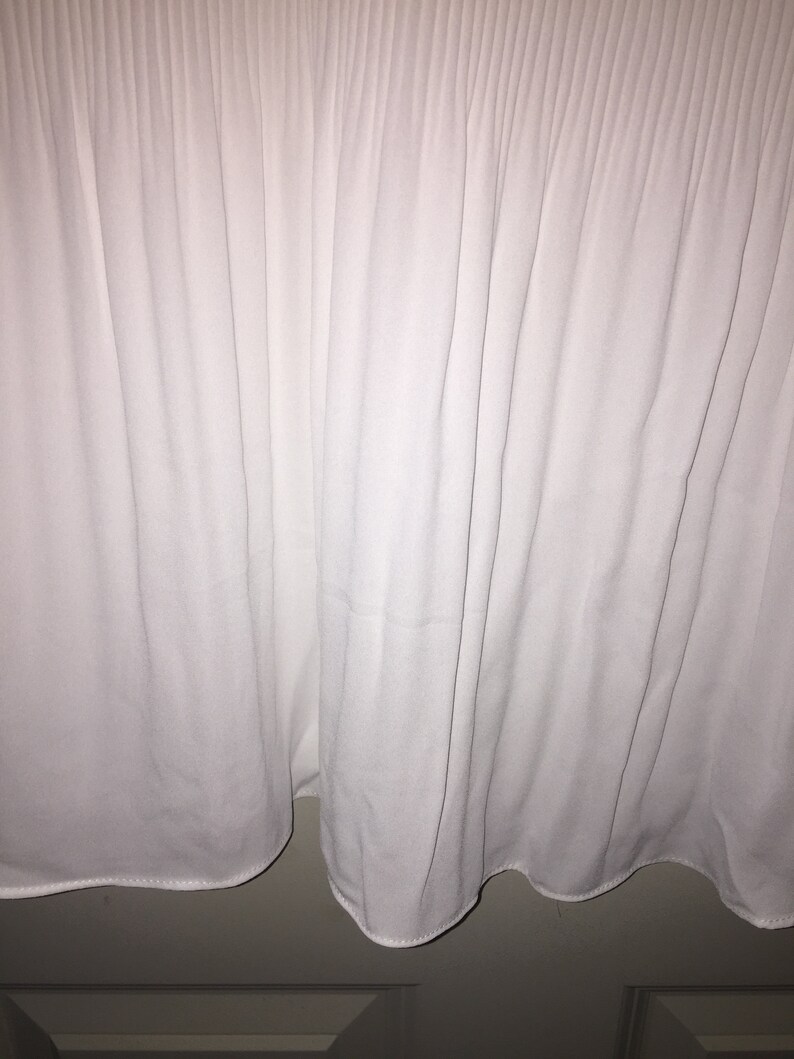 May include: White pleated fabric with a smooth texture. The fabric is gathered at the bottom, creating a ruffled edge.