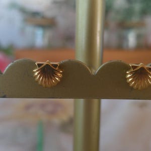 May include: A pair of gold-colored seashell stud earrings. The earrings are displayed on a gold-colored jewelry stand. The earrings have a detailed, textured design, and are small in size.