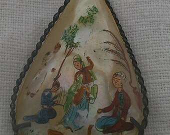 May include: A teardrop-shaped pendant with a silver-toned metal frame and a painted scene of three figures in traditional clothing. The figures are surrounded by green and brown foliage. The pendant is a vintage piece of jewelry.