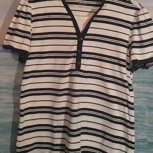 May include: A short-sleeved, striped shirt in white and navy blue. The shirt has a V-neck with a button-down placket and black trim on the collar and sleeves. The brand name "Van Heusen" is visible on the label.