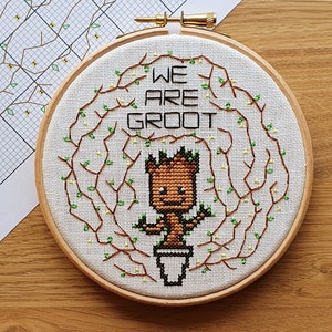 May include: A cross-stitch embroidery hoop with a white background and brown branches with green leaves. The center of the hoop features a cartoon image of a brown and green character with the text "We Are Groot" above it.
