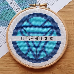 May include: A cross-stitch embroidery in a wooden hoop. The design features a blue and teal geometric pattern with the text "I LOVE YOU 3000" in white. The background is a light-colored fabric.