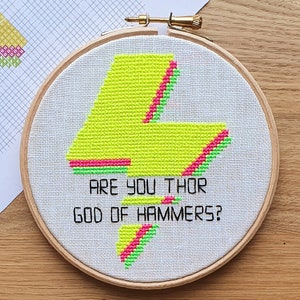 Può includere: Un cerchio da ricamo bianco con un fulmine giallo e rosa neon e il testo "Are you Thor God of Hammers?"
