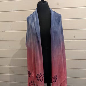 May include: A blue and pink ombre shawl with paw print accents. The shawl is displayed on a black mannequin. The color gradient transitions from blue at the top to pink at the bottom. Dark blue paw prints are along the bottom edge.