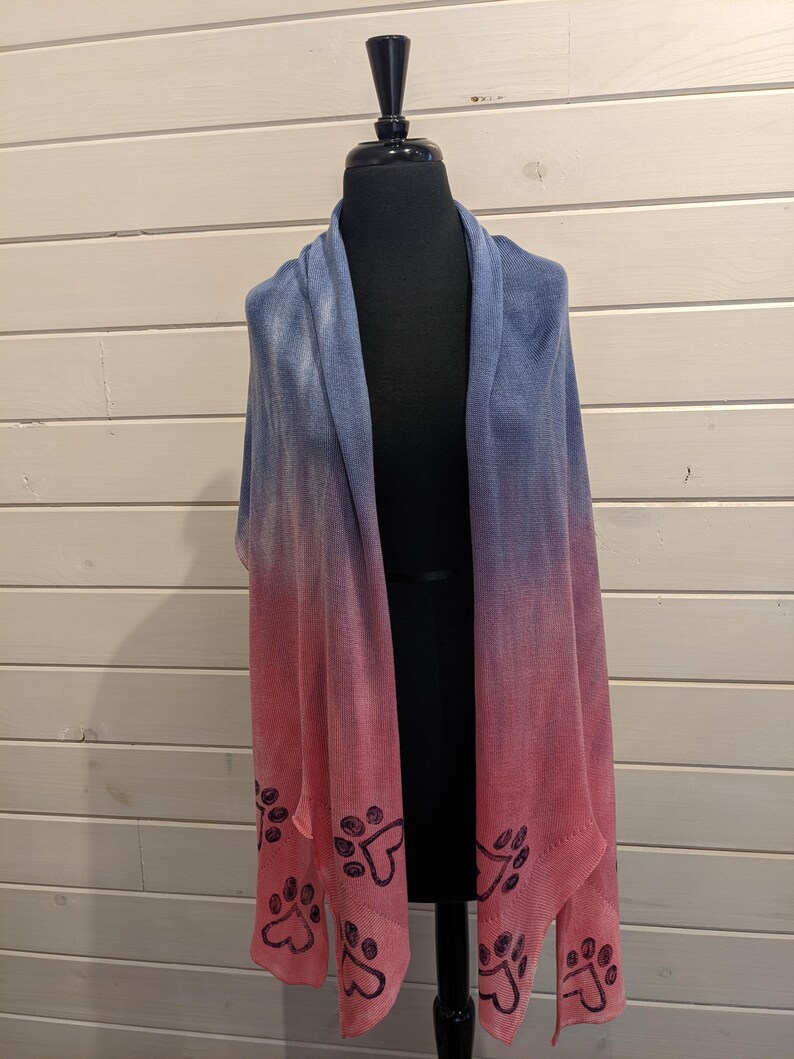 May include: A blue and pink ombre shawl with paw print accents. The shawl is displayed on a black mannequin. The color gradient transitions from blue at the top to pink at the bottom. Dark blue paw prints are along the bottom edge.