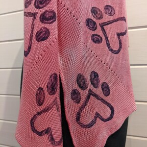 May include: A pink knitted scarf featuring a repeating pattern of dark purple paw prints with heart-shaped pads. The scarf is draped, showcasing its triangular shape and textured knit. The background is a light-colored wooden surface.