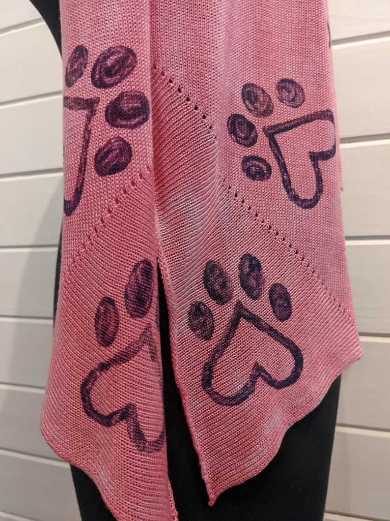 May include: A pink knitted scarf featuring a repeating pattern of dark purple paw prints with heart-shaped pads. The scarf is draped, showcasing its triangular shape and textured knit. The background is a light-colored wooden surface.