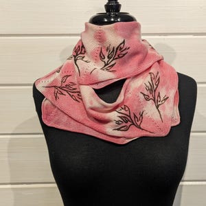 May include: A pink and white knitted scarf with a leaf design. The scarf is displayed on a black mannequin. The leaf design is in black. The background is a white wooden wall.