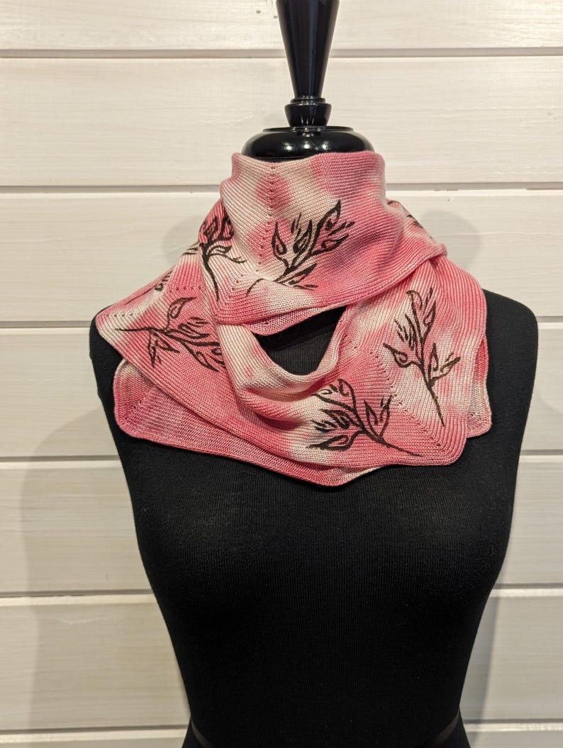 May include: A pink and white knitted scarf with a leaf design. The scarf is displayed on a black mannequin. The leaf design is in black. The background is a white wooden wall.