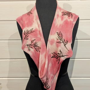 May include: A pink and white tie-dye knit cowl scarf with a leaf design. The scarf is displayed on a black mannequin. The leaf design is in a dark brown color. The scarf is a soft, lightweight accessory.